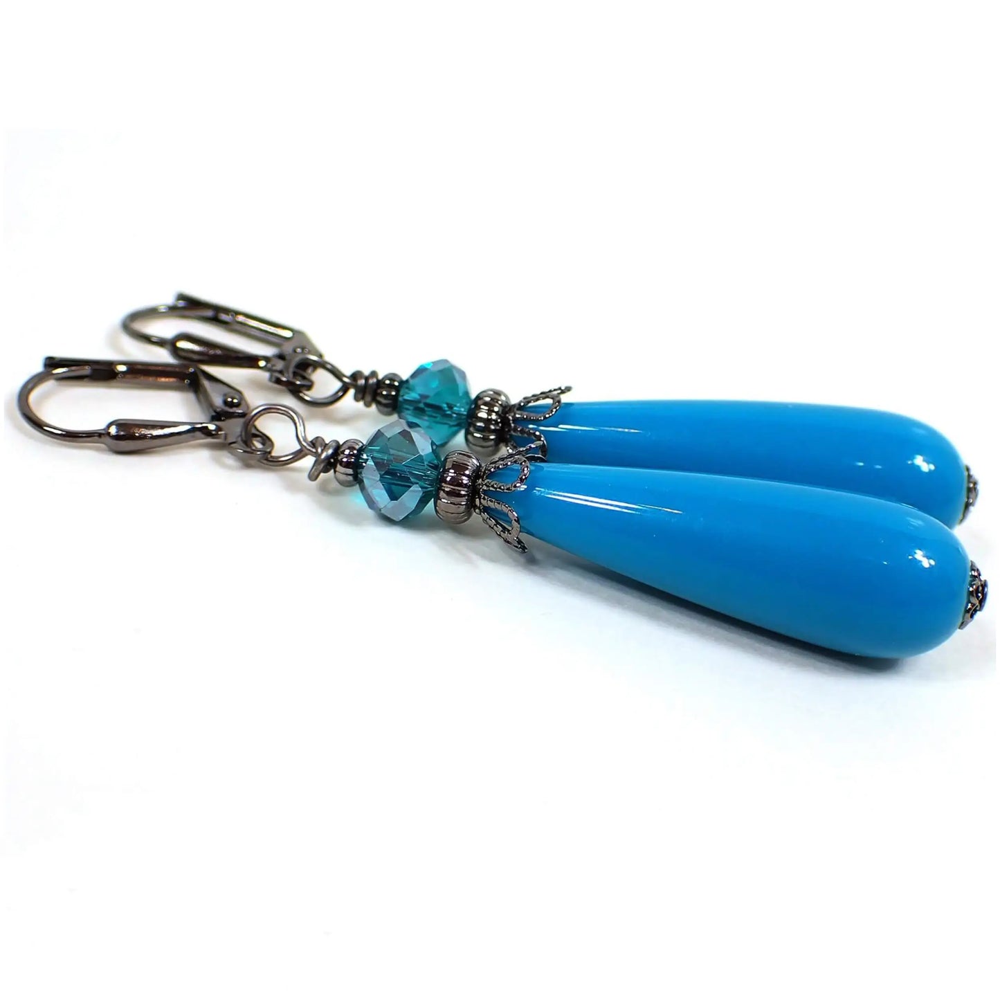 Angled view of the handmade teardrop earrings with vintage lucite beads. The metal is gunmetal gray in color. There are teal blue faceted glass crystal beads at the top. The bottom lucite beads are teardrop shaped and a dark teal blue in color.