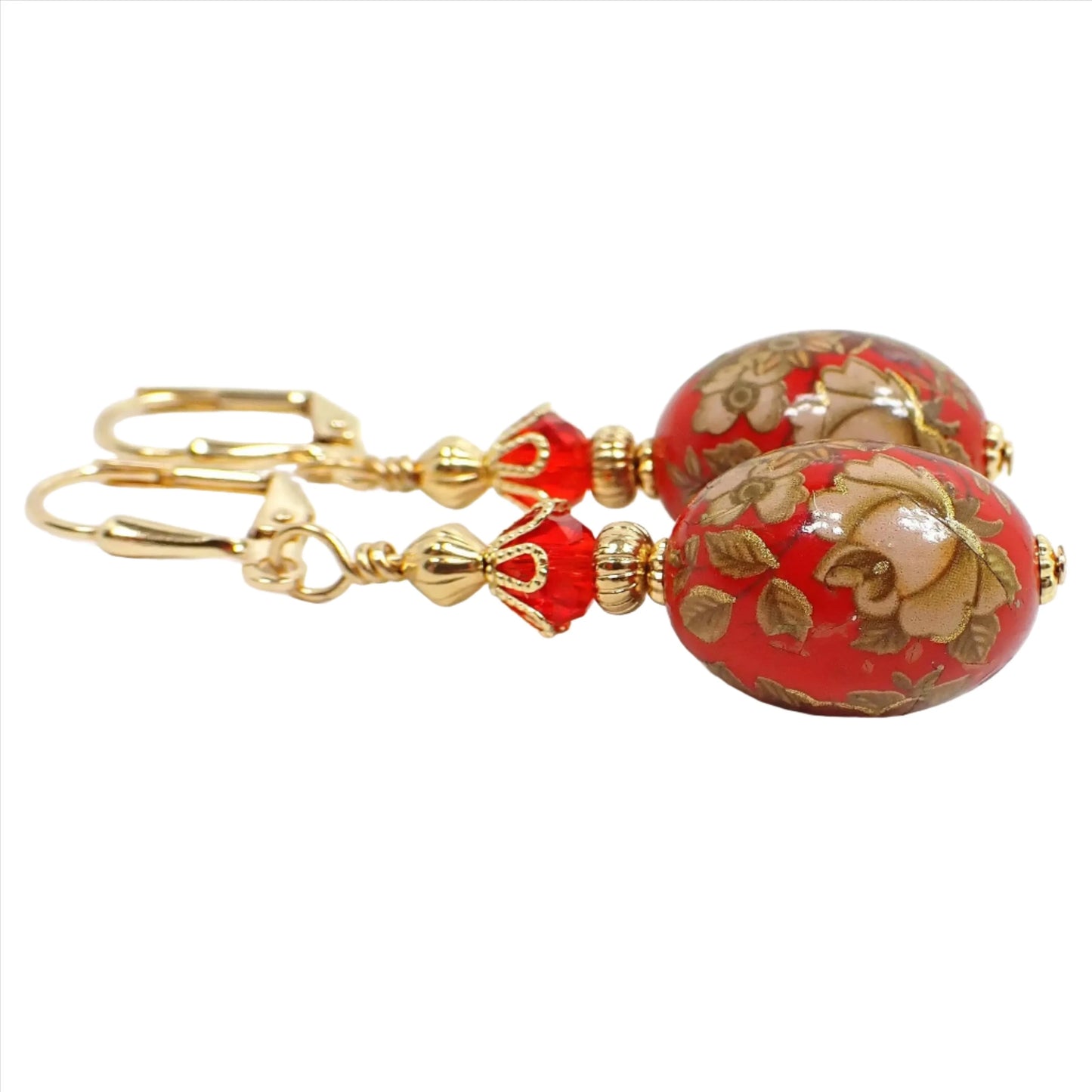 Side view of the handmade floral drop earrings. The metal is gold plated in color. There are bright red faceted glass crystal beads at the top. The bottom acrylic beads are oval shaped, deep red in color, and have flower decals around them. The decals have flowers with off white and brown tones and metallic gold edging.