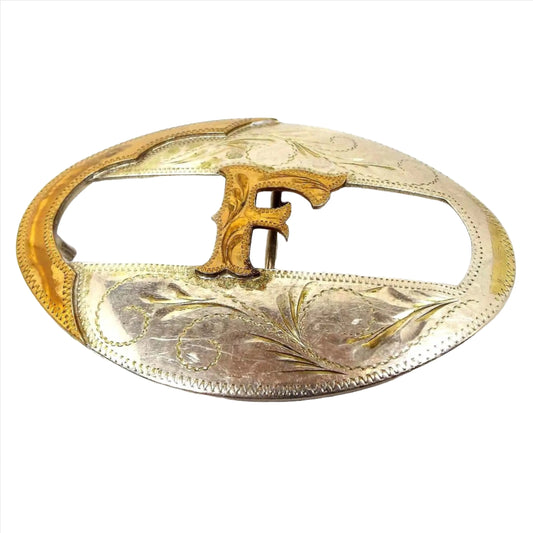 Front view of the retro vintage initial letter F belt buckle. It is mostly silver tone in color with brass edges and a brass letter F in the middle. The middle area has a cut out design and there is a leaf and floral pattern stamped on the buckle.
