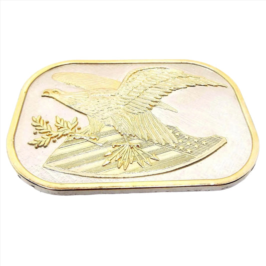 Angled front view of the retro vintage flying eagle and shield belt buckle. The majority of the belt buckle is silver tone in color. The design is in gold tone color. There is a flying eagle holding a branch and arrows over a shield with the stars and stripes on it.