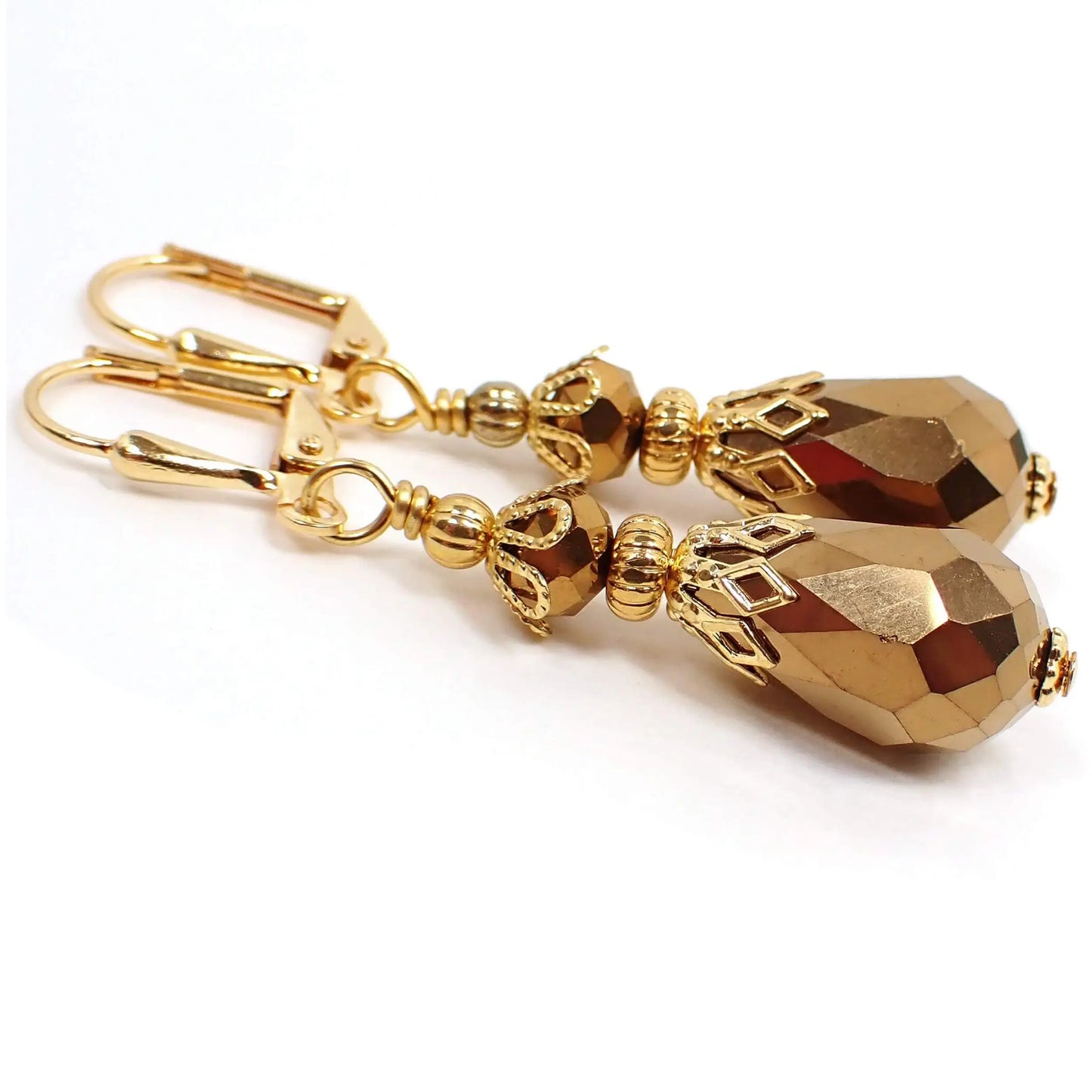 Angled view of the handmade metallic teardrop earrings. The metal is gold plated in color. The beads are faceted glass crystal and a metallic bronze in color. The top bead is rounded saucer shaped and the bottom bead is a teardrop.