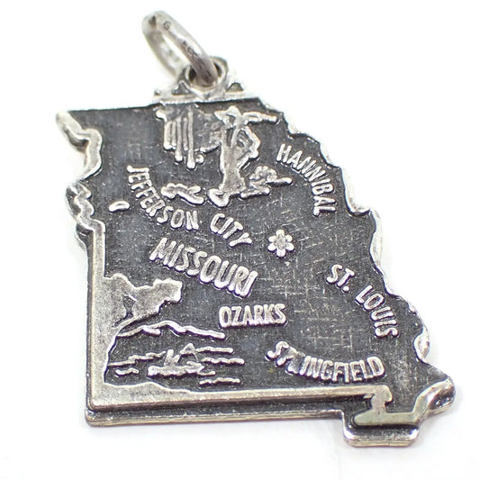 Enlarged view of the retro vintage sterling silver souvenir charm. It's shaped like the state of Missouri and has several cities on it, as well as depictions of notable things. The sterling has darkened for an antiqued silver color of silver and dark gray. There is a small jump ring attached to the top.