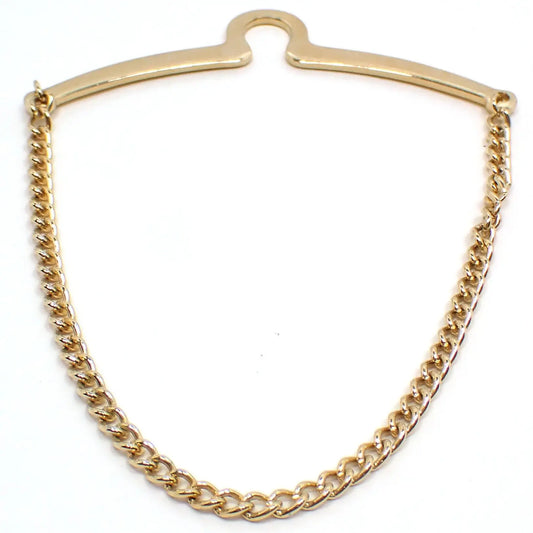 Enlarged front view of the retro vintage tie chain. The metal is gold tone plated in color. There is a curved bar at the top with a curve in the middle to go over a button. The chain is curb link and hangs down from one side to the other.