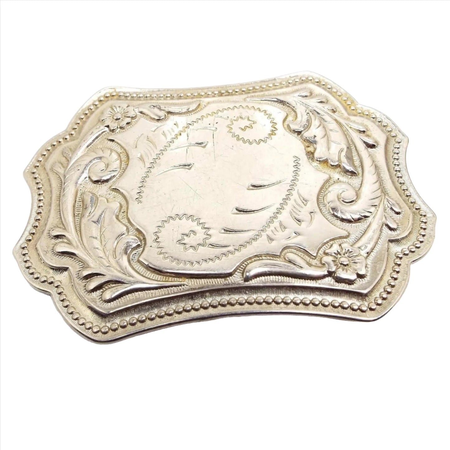 Front view of the retro vintage silver tone belt buckle. It has an etched and raised curled leaf like design and a dotted pattern around the edge. There is a flower on two of the corners. A few small scuff scratches from age can be seen on the front.