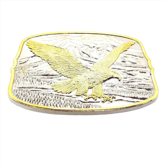 Front view of the retro vintage flying eagle belt buckle. It has an eagle flying over trees by mountains design. The metal is mostly silver tone in color with a gold tone edge and gold tone color on the eagle as well.