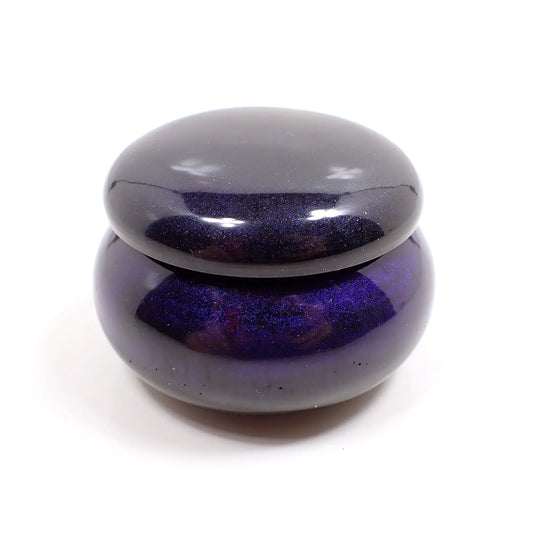 Side view of the small handmade round trinket box jar with lid. It is dark in color with hints of dark pearly purple and blue depending on how the light hits it.