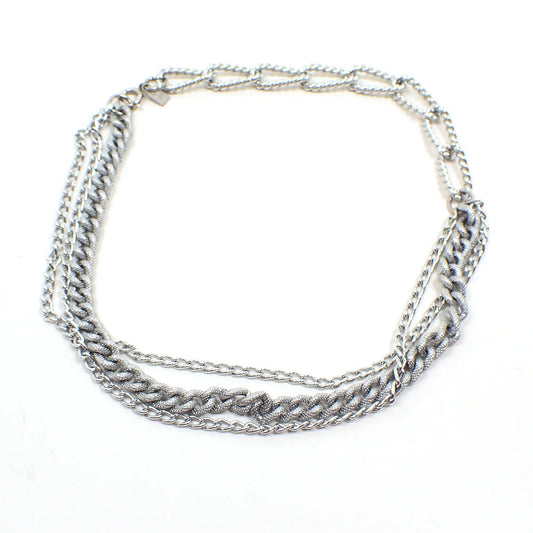 Top view of the retro vintage Emmons multi strand chain necklace. The metal is silver tone in color and has a spring ring clasp at the end. Part of the necklace has larger sized textured oval curb links and the other part has three strands of small sized link chains. Two of the three strands are shiny and the other is slightly bigger with textured links.