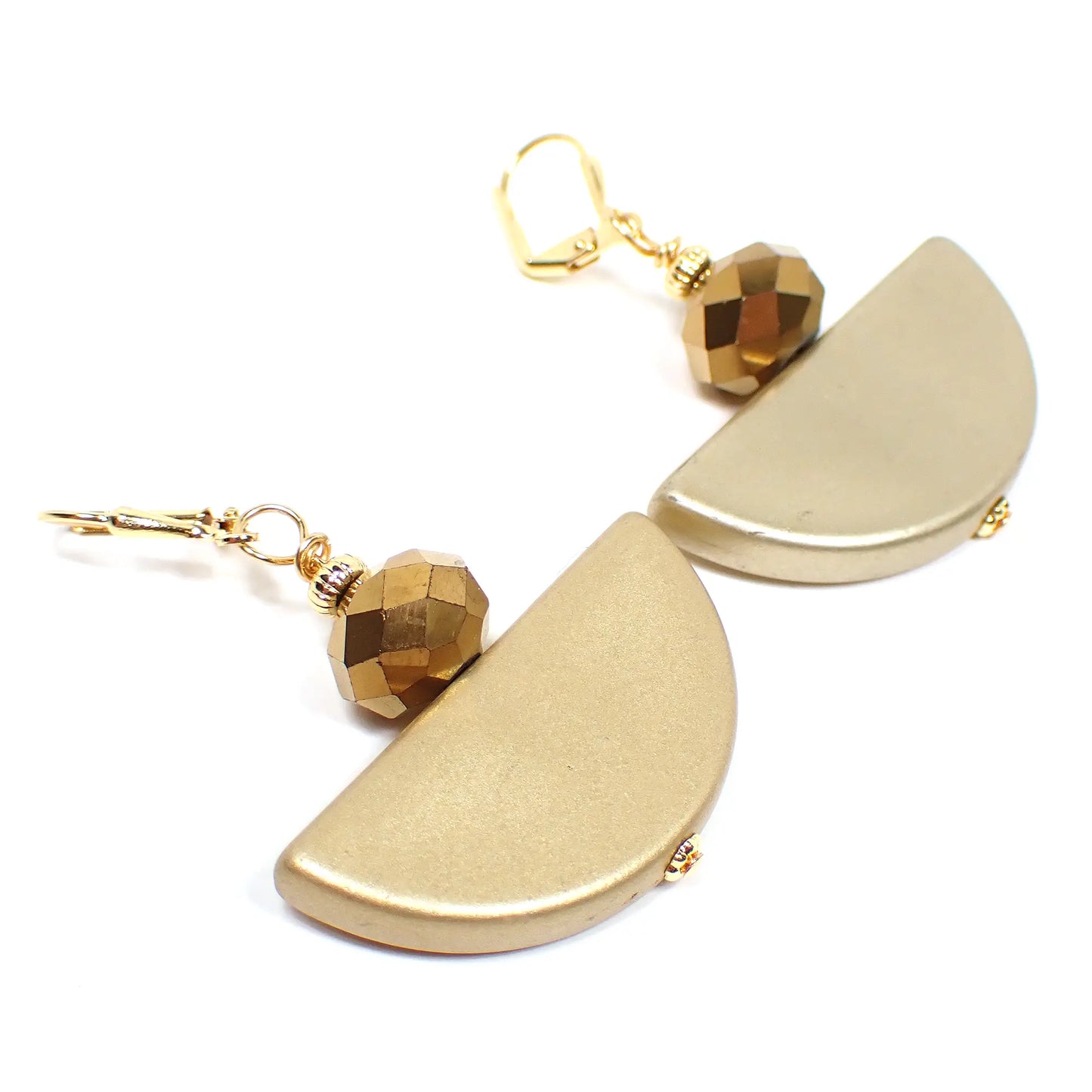 Angled view of the handmade half moon earrings. The metal is gold plated in color. There are faceted round antiqued metallic gold colored glass beads on top. The bottom beads are vintage acrylic beads that are half moon shaped and a lighter matte metallic gold in color.
