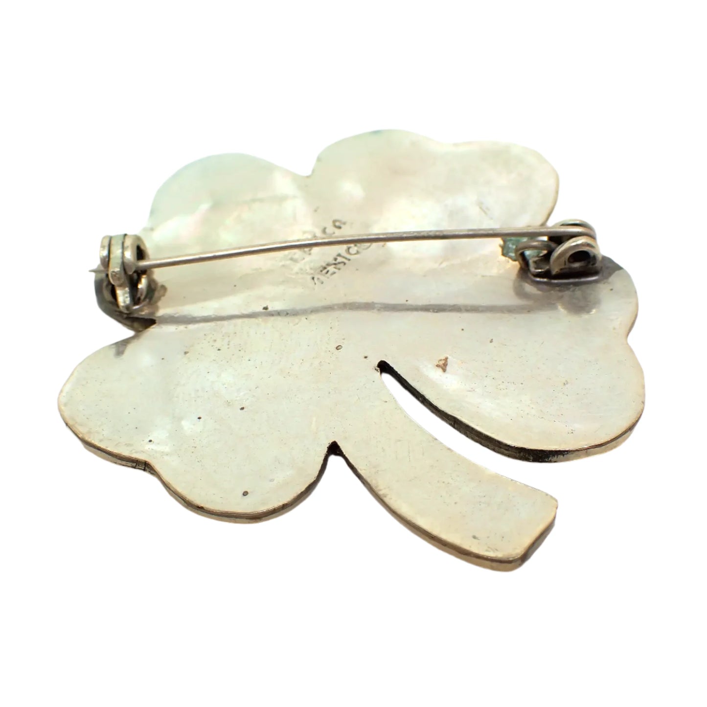 Photo of the back of the four leaf clover brooch. The metal is darkened and a small green spot of verdigris can be seen on the right by the pink hinge. The pin is slightly bent but still holding in the clasp.