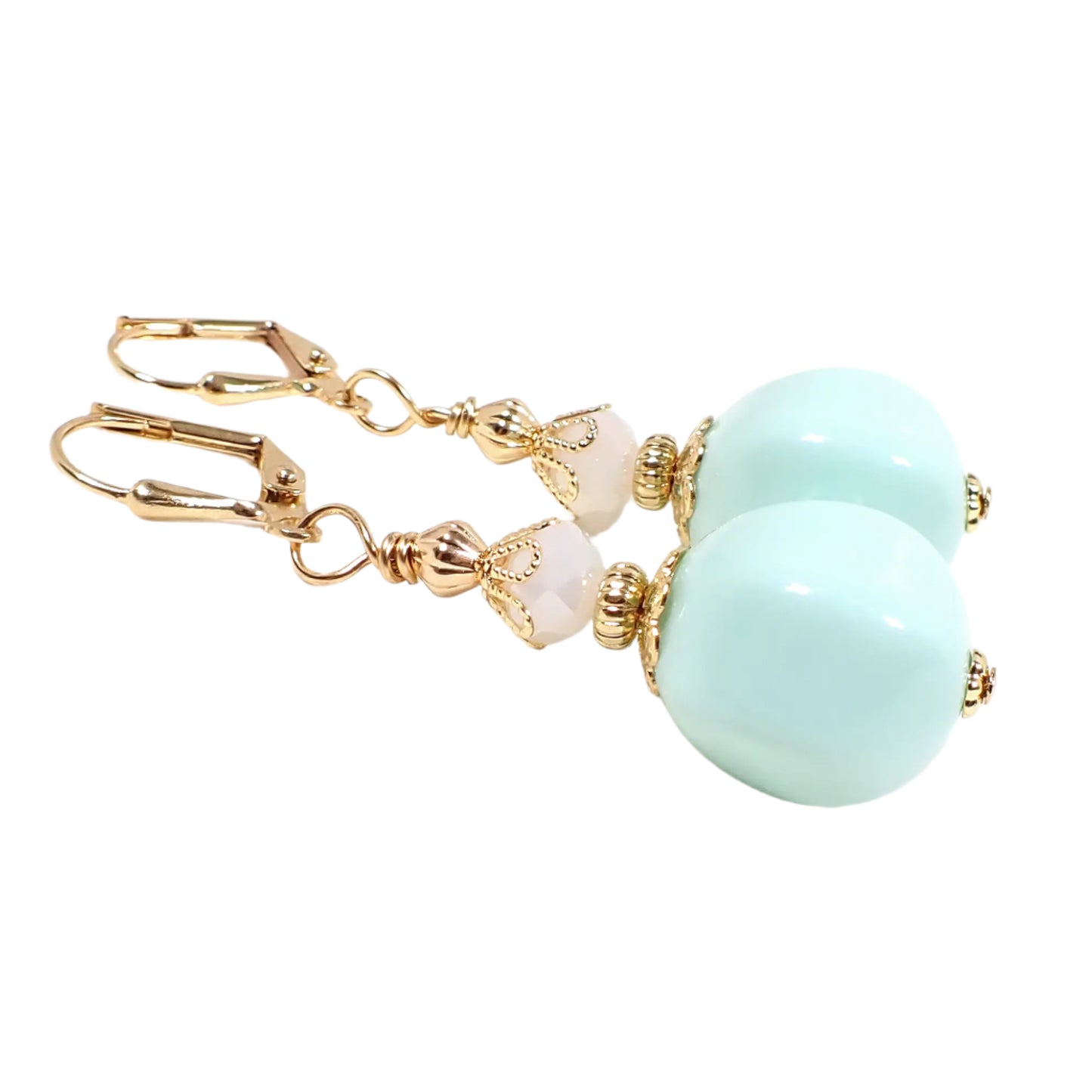 Angled view of the handmade beaded drop earrings. The metal is gold tone in color. There are faceted white glass beads at the top. There are vintage German acrylic beads at the bottom in an opaque light green color. The acrylic beads have a nugget like shape.