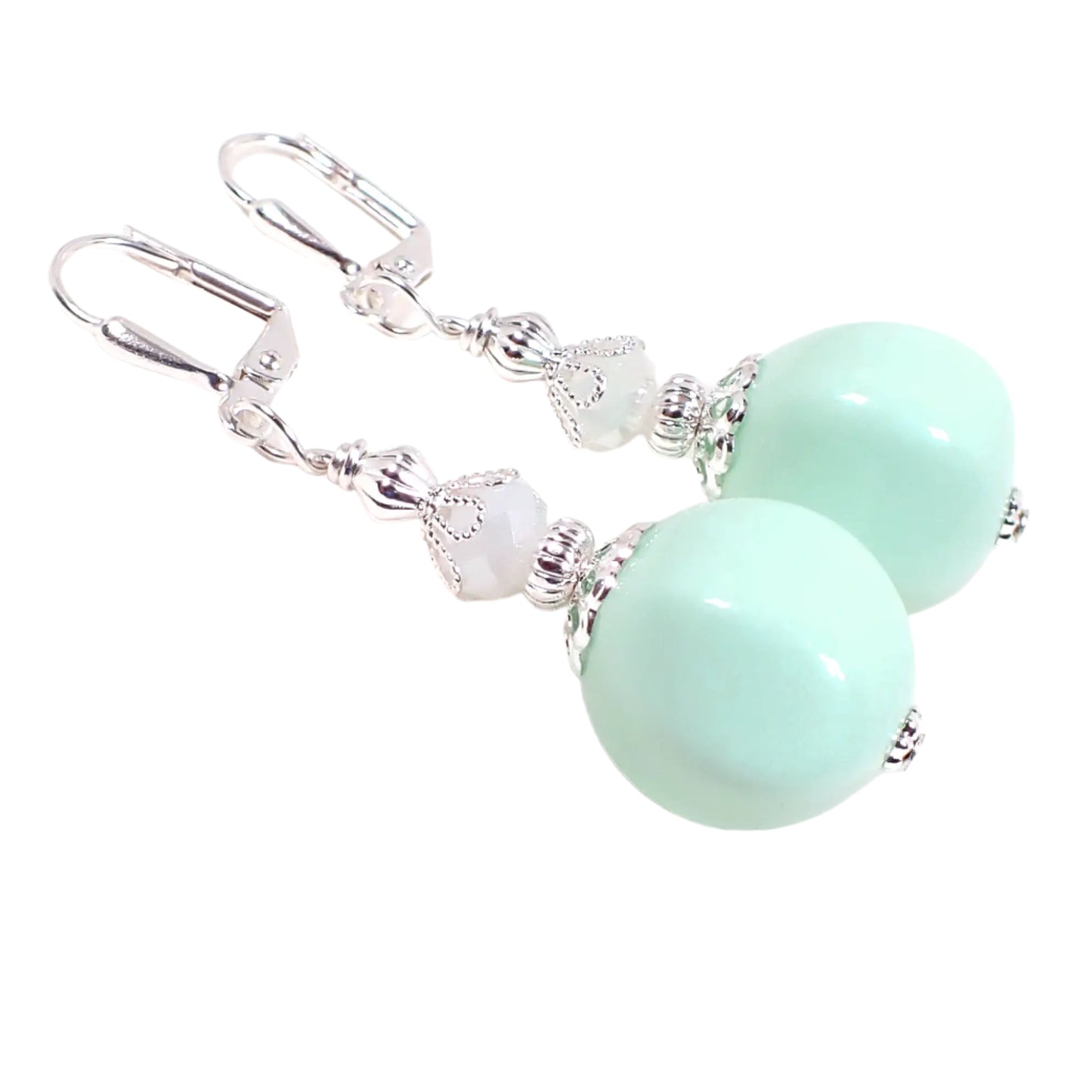 Angled view of the handmade beaded drop earrings. The metal is silver tone in color. There are faceted white glass beads at the top. There are vintage German acrylic beads at the bottom in an opaque light green color. The acrylic beads have a nugget like shape.