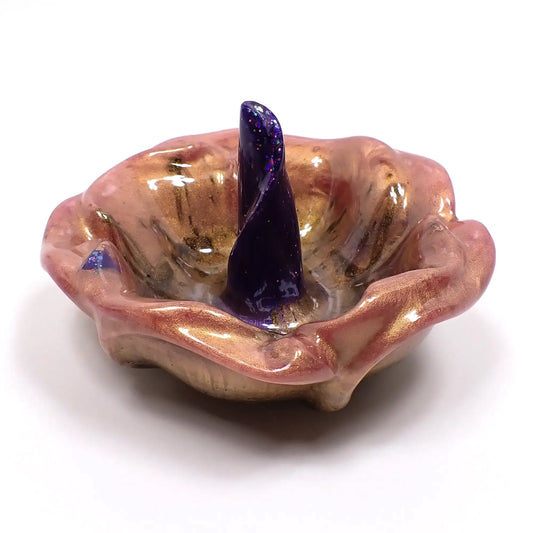 Angled side view of the handmade resin ring dish. It is shaped like an open flower with petals around the edge. There is a area in the middle shaped like a curled petal that is standing straight up for rings to slide over and sit upon. The outer petals are marbled with shades of pink, light purple, and brown. The inside petal has shades of vivid purple and blue with iridescent glitter.