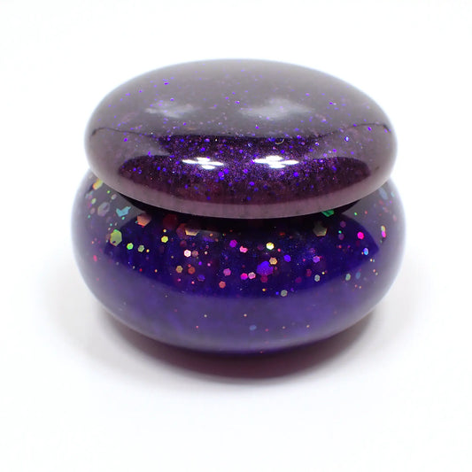 Side view of the small handmade resin trinket box. It has a rounded short jar like shape. The lid has pearly dark purple resin with tiny flecks of blue glitter. The bottom has a vivid pearly blue resin with chunky iridescent purple glitter.