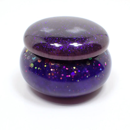 Side view of the small handmade resin trinket box. It has a rounded short jar like shape. The lid has pearly dark purple resin with tiny flecks of blue glitter. The bottom has a vivid pearly blue resin with chunky iridescent purple glitter.