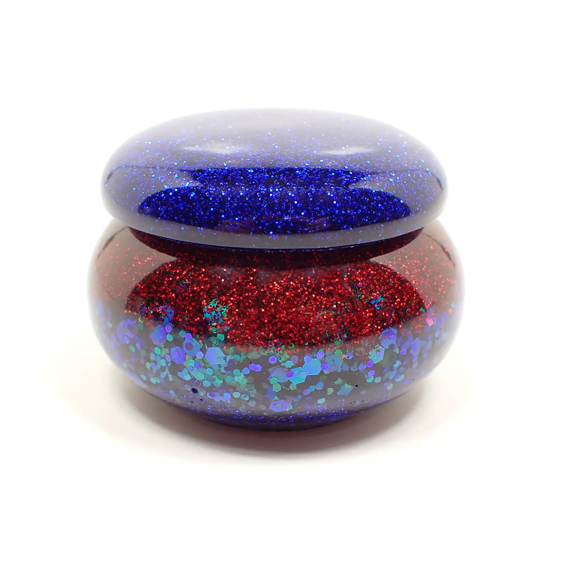 Side view of the handmade small resin trinket box. It has a short round jar like shape. The lid is pearly blue with blue glitter. The bottom part has a band of red with red glitter, a band of chunky iridescent green glitter, and then a small band of blue with blue glitter at the bottom. Very sparkly.