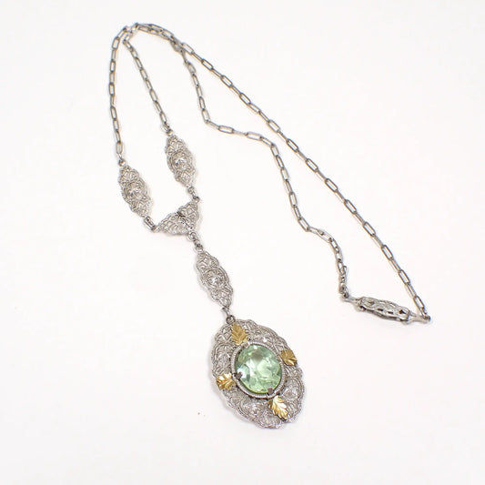 Front view of the 1920's Art Deco vintage rhodium plated pendant necklace. The metal is silver tone in color. There are square flat cable links on the chain with a clip clasp. The bottom part of the necklace has filigree links and oval pendant drop with gold plated leaf accents and a faceted light green oval Czech glass cab.