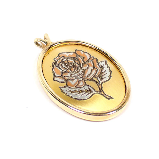 Angled view of the retro vintage Reed and Barton pendant. It is oval in shape. There is an etched rose flower design on the front with copper and silver color accents. The rest of the pendant is gold tone in color. There is a double opening V shaped bail at the top. There is no chain.