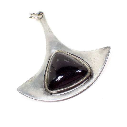 Angled view of the Mid Century vintage Corocraft pendant. The metal is matte silver tone in color. it has a long thin top that flares out at the bottom. There is a domed rounded triangle black glass cab in the middle. A couple of tiny spots of discoloration can be seen on the left under magnification. There is no chain.