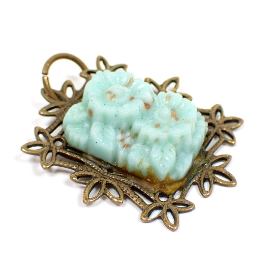 Angled view of the Art Deco Czech glass pendant. The metal is brass and has a stamped filigree design with leaf like edge. There is a Czech molded glass cab in the middle with a design of two flowers and leaves in a mint green color with specks of brown. The adhesive around the edge has yellowed with age and a thin line of verdigris can be seen on the bottom.