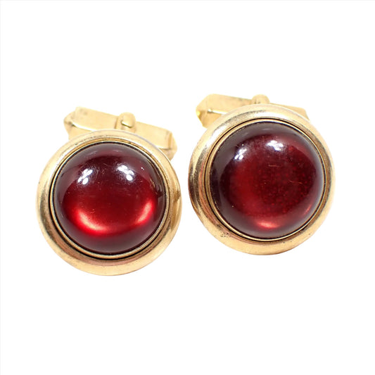 Front view of the Swank Mid Century vintage cufflinks. The metal is gold tone in color. They are round with maroon red domed moonglow lucite cabs on the front. The backs are angled.