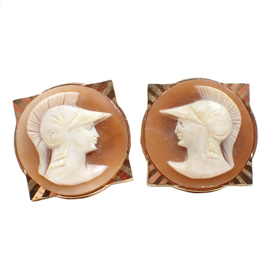Front view of the Mid Century vintage Swank cameo cufflinks. The metal is gold tone in color and has a square shape with diamond cut diagonal lines. On the front of that is a round area with a carved shell cameo depicting the Greek Goddess Athena. The background area of the shell is peach in color and the cameo part is mostly white in color.