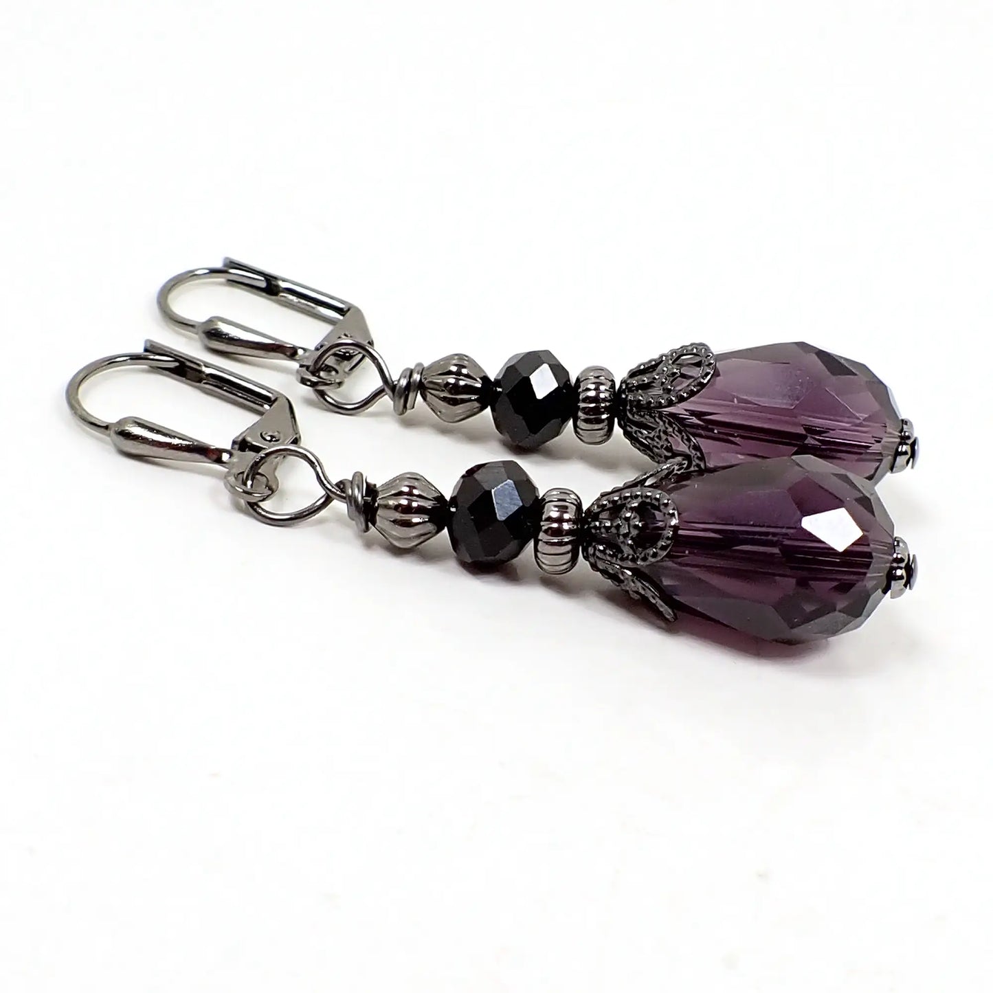 Angled view of the handmade drop earrings. The metal is gunmetal dark gray in color. There is a faceted black glass crystal saucer bead at the top and a faceted dark purple glass crystal teardrop bead at the bottom.
