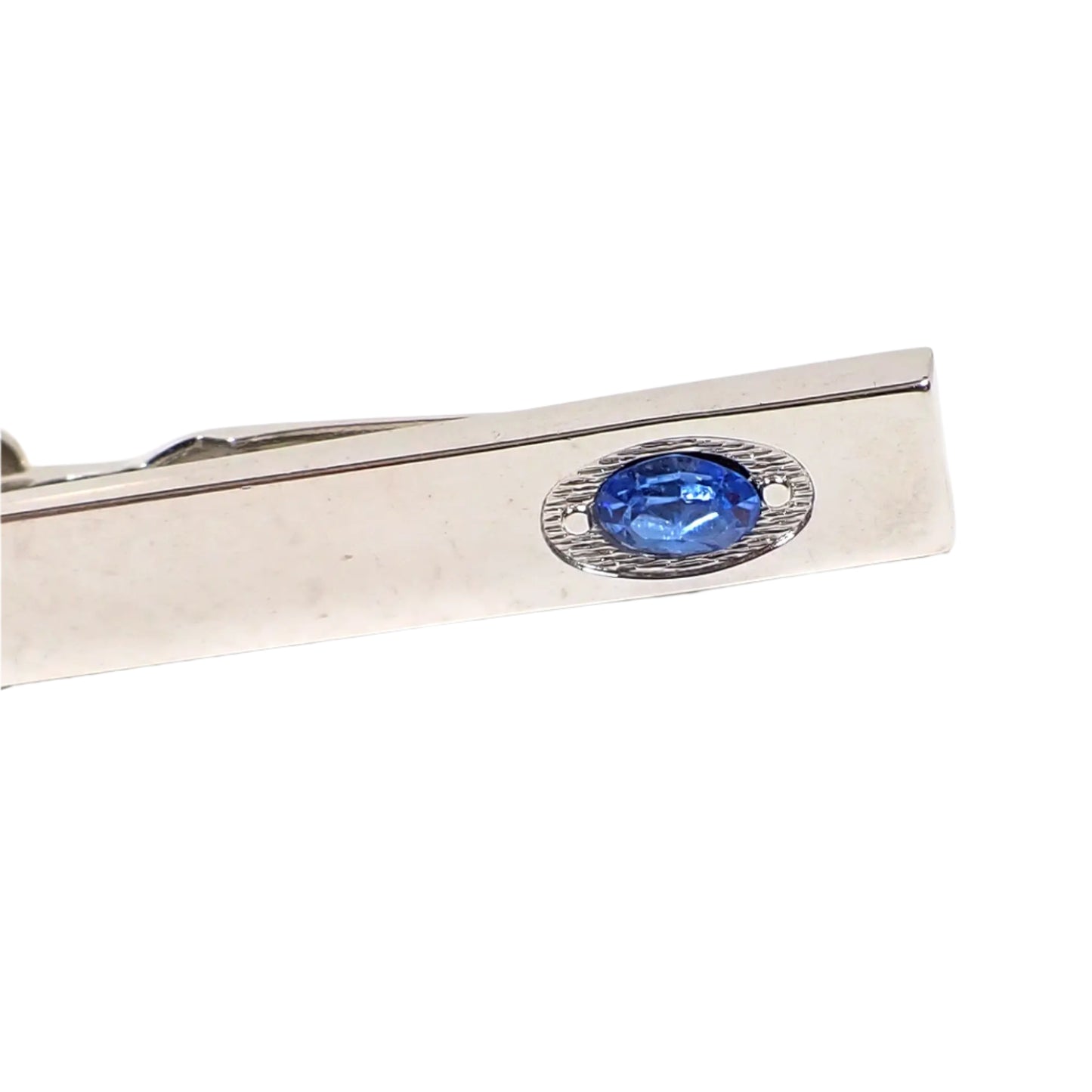 Closeup view of the blue rhinestone on the end of the vintage Swank tie clip. There is and indented oval area at the end of the tie clip that has a textured small wavy line pattern. There is a shiny raised flat dot on the sides of the rhinestone. The rhinestone is also oval shaped.