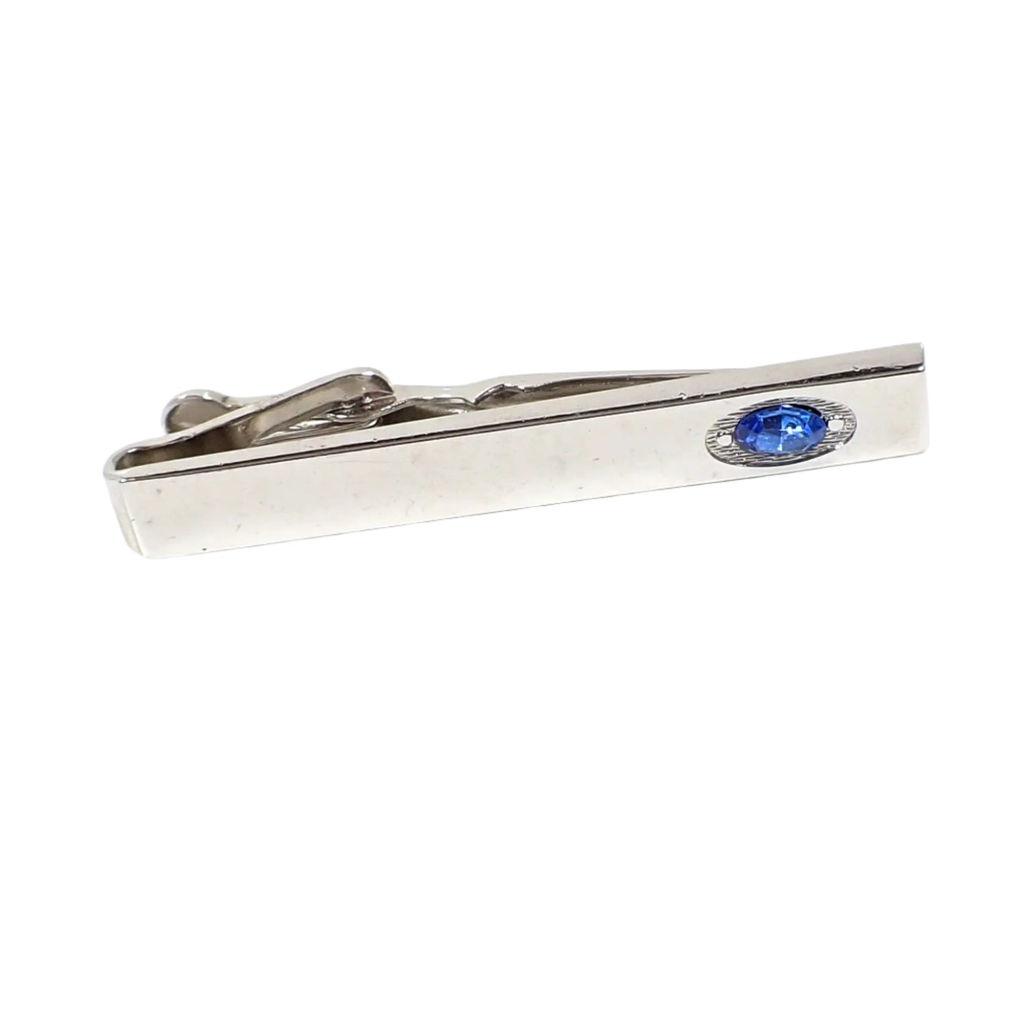 Angled view of the vintage Swank tie clip. The metal is shiny silver tone in color and there is an oval blue rhinestone on the end. Part of the alligator style clip on the back is showing in the photo.