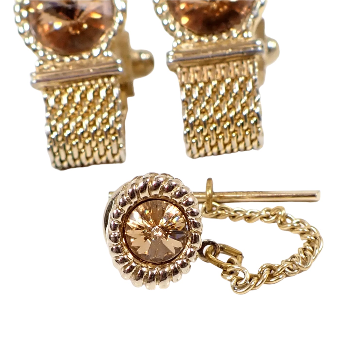 Close up view of the tie tack. It is oval with a braided rope style metal design around the rivoli rhinestone. The rhinestone is round and sparkly in a champagne color. The clutch on the back has a chain with a bar on the end.