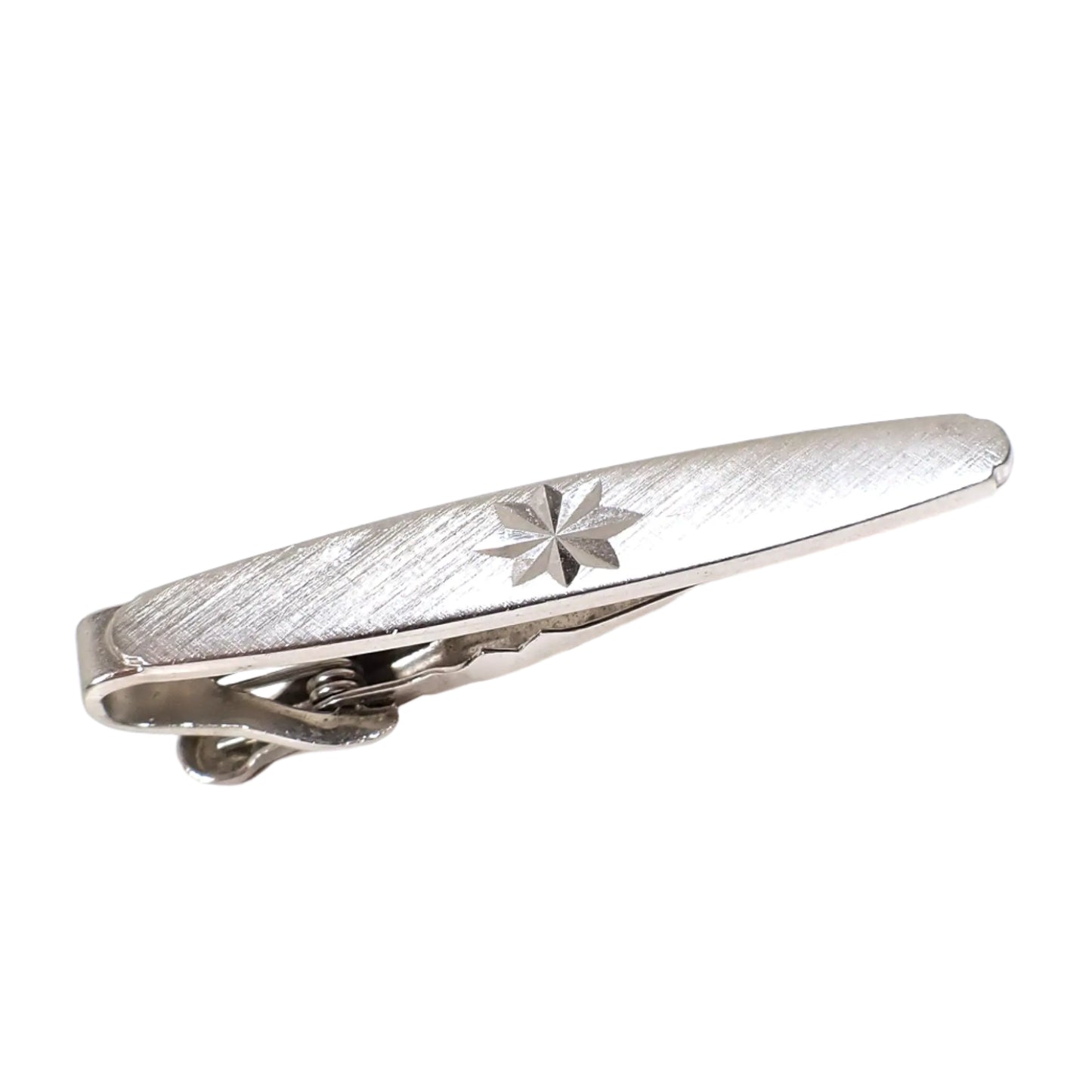 Angled top view of the Swank vintage tie clip. The metal is silver tone in color. It is oval shaped with fancy ends and has a matte brush textured front. There is a starburst design etched in the middle of the tie clip.