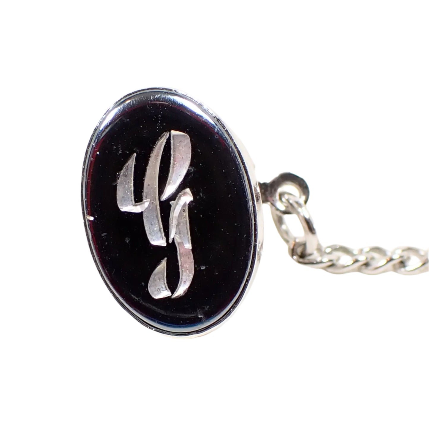 Front view of the Mid Century vintage faux hematite tie tack. The metal is silver tone in color. The tie tack is oval and has a dark metallic gray imitation hemtatite front with a fancy script initial G in the middle.