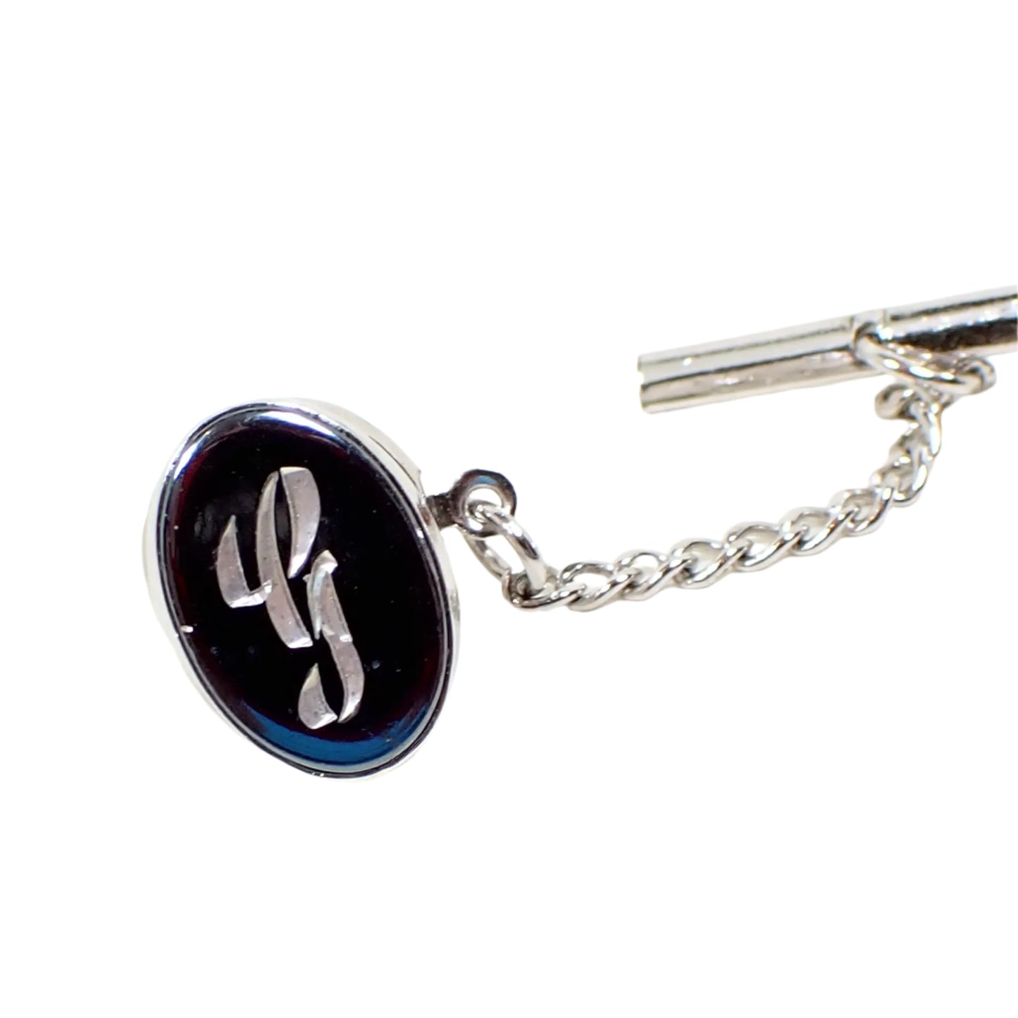 Full view of the vintage initial tie tack. It is oval shaped with silver tone color metal and a dark metallic gray faux hematite front. There is a fancy script letter G engraved in the middle.