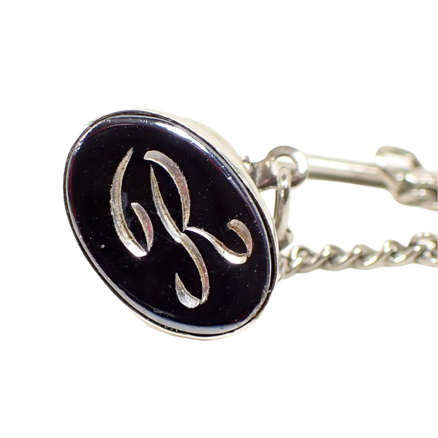 Front view of the Mid Century vintage faux hematite tie tack. The metal is silver tone in color. The tie tack is oval and has a dark metallic gray imitation hemtatite front with a fancy script initial R in the middle.