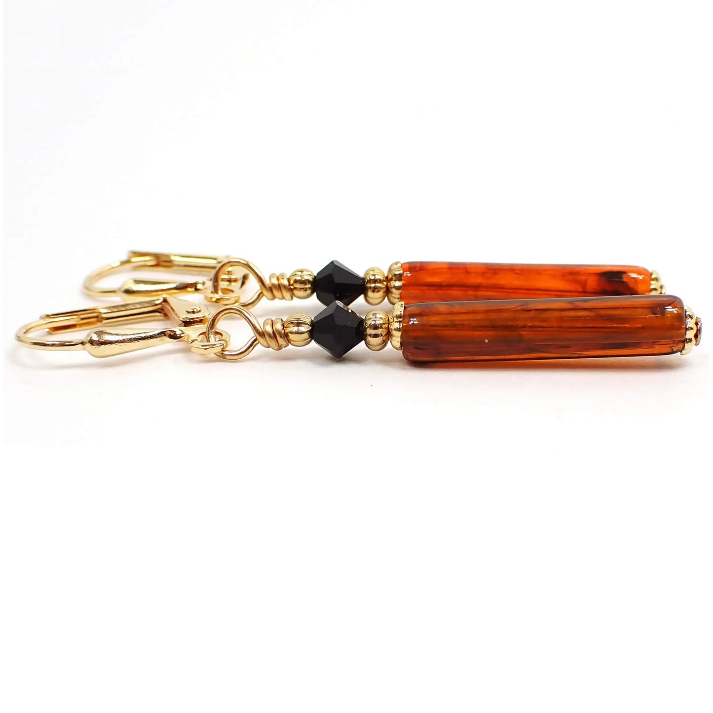 Side view of the handmade faux tortoise stick earrings. The metal is gold plated in color. There are faceted black glass crystal beads at the top. The bottom lucite beads are tube shaped stick like and have shades of orange, brown, and black in them.