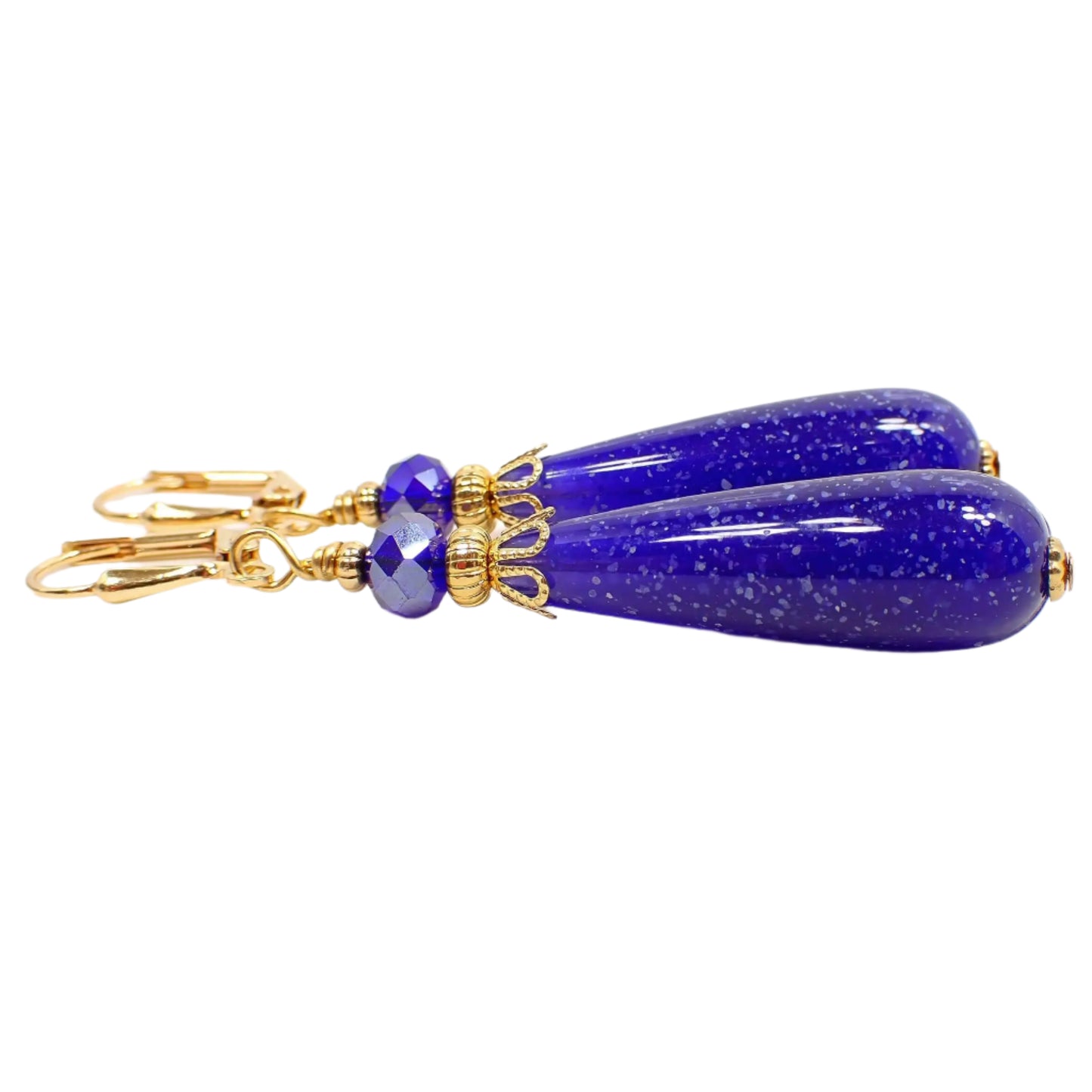Side view of the handmade teardrop earrings. The metal is gold plated in color. There is a faceted cobalt blue glass crystal bead at the top. The bottom bead is a rounded teardrop shape with dark blue lucite and small flecks of white confetti. The confetti lucite beads are vintage beads that were never before used.
