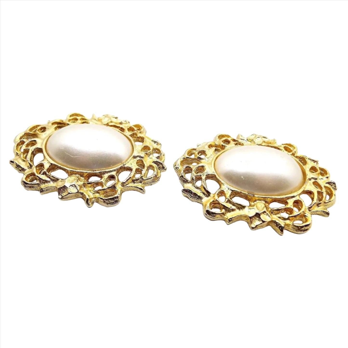 Front view of the retro vintage Bluette vintage shoe clips from the 1980's. The middle areas have large rounded oval pearly white plastic faux pearl cabs. Surrounding the imitation pearl areas is a wreath like filigree design that goes all the way around the edge. The metal is gold tone in color.