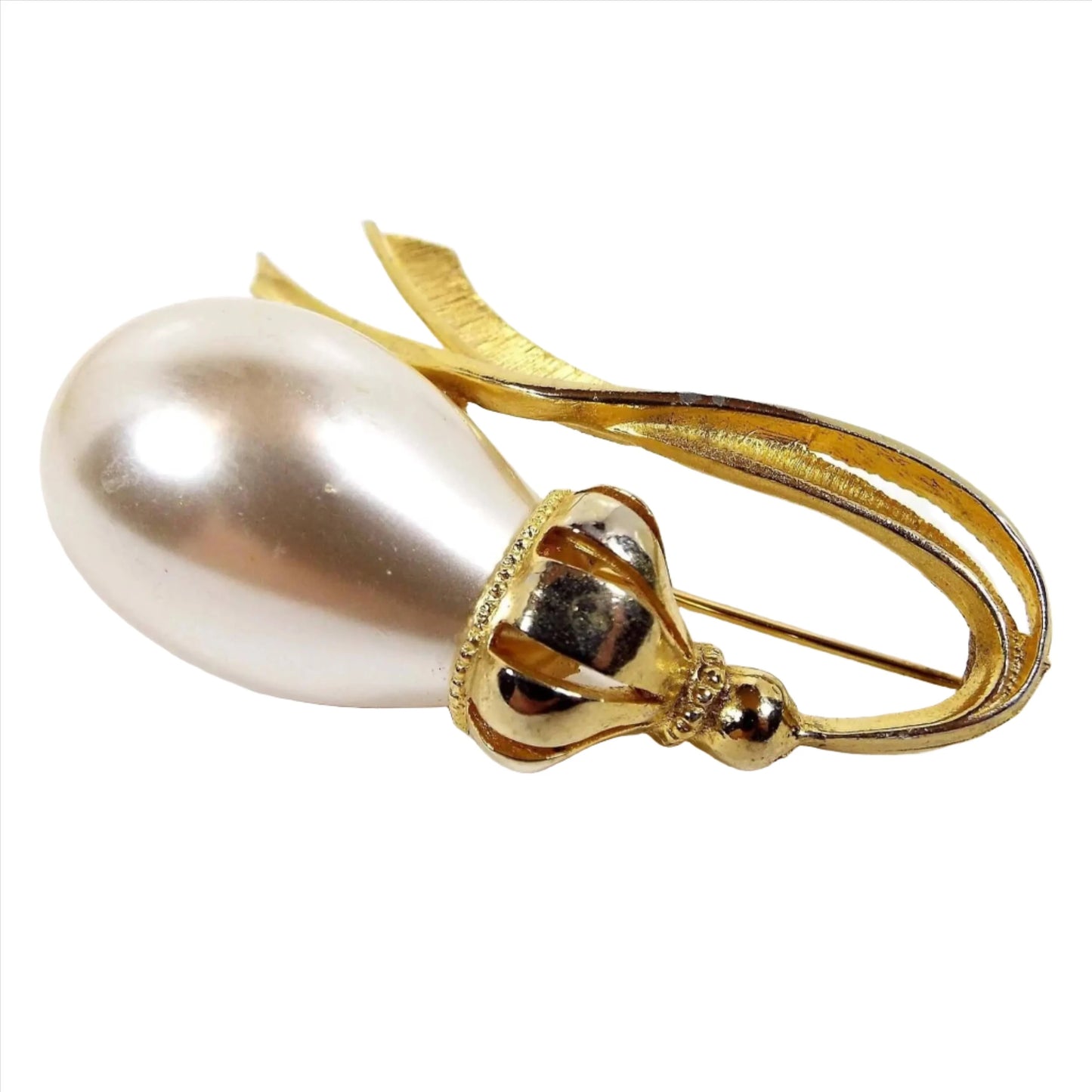 Front view of the retro vintage faux pearl brooch. The metal is gold tone in color. There is a large off white coated plastic rounded teardrop imitation pearl on the left side. Past that is a bulbous area with cut out lines down to a round ball shape and then metal shaped like two ribbons coming off the very end and curving back around to the faux pearl.