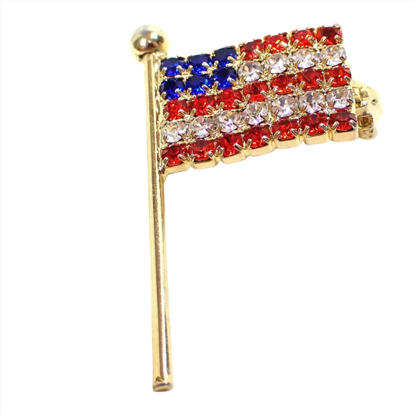Enlarged front view of the retro vintage rhinestone flag pin. The metal is gold tone plated in color. The rhinestones are red white and blue like a US flag.