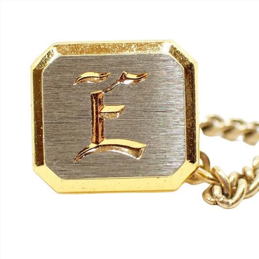 Front view of the Mid Century vintage initial tie tack. Most of the tie tack is gold tone in color including the back, edge, and the engraved letter itself. The front middle area is silver tone in color with a matte appearance. The tie tack is shaped like an octagon and has the letter E engraved on the front.