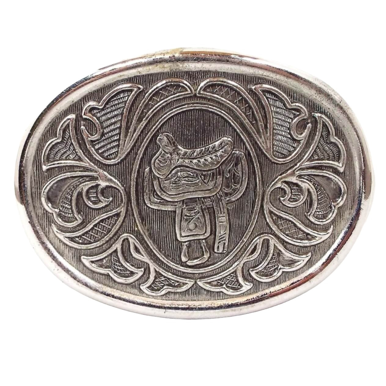 Front view of the retro vintage Avon belt buckle. It is silver tone in color with a light gray background. There is a horse saddle in the middle and a floral like pattern around the edge.