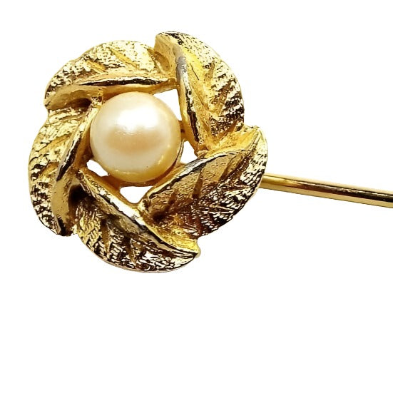 Enlarged top view of the Mid Century vintage faux pearl stick pin. The metal is gold tone in color. The top has a wreath style design made of textured metal leaves. There is an off white imitation pearl in the middle.