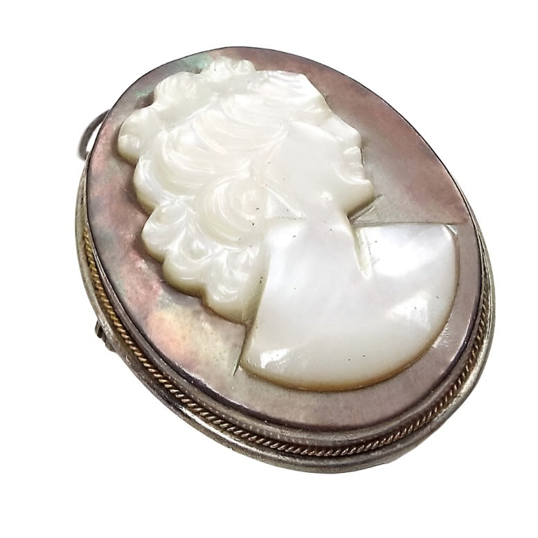 Angled front view of the 1940's vintage cameo brooch pendant. It is oval in shape with mostly a silver tone color bezel setting that has a thin twisted style gold tone trim around the edge. The middle is a carved mother of pearl cab. The middle part is a pearly white bust of a woman and the background is a pearly brownish gray in color.