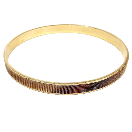 Angled view of the retro vintage gold tone bangle bracelet. The edge has small facets and there is a etched cut diagonal line pattern all the way around