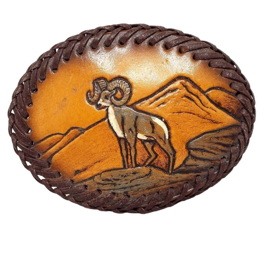 Front view of the retro vintage goat ram vintage belt buckle. It is covered with leather and has a leather cord sewn around the edge. In the middle is a ram standing on a rock in front of some mountains. The leather is dyed in shades of brown and orange.