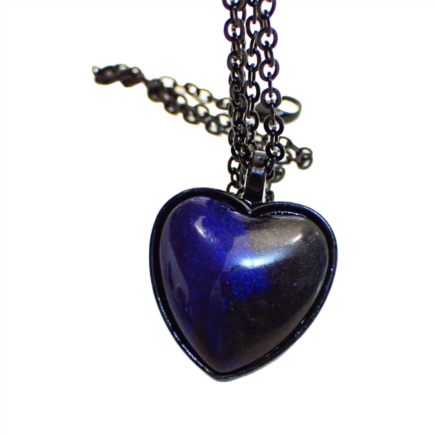 Enlarged front view of the Goth black heart handmade pendant necklace. The chain and pendant setting are black coated. The pendant is heart shaped with a domed handmade resin cab. The resin is pearly dark blue and black in color.