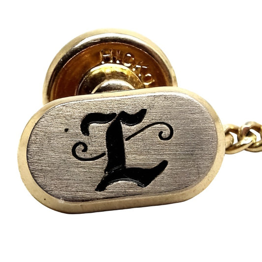 Enlarged front view of the Mid Century vintage Hickok initial tie tack. The front of the oval tie tack is silver tone in color with a black fancy script letter L on the front. The rest of the tie tack is gold tone in color.
