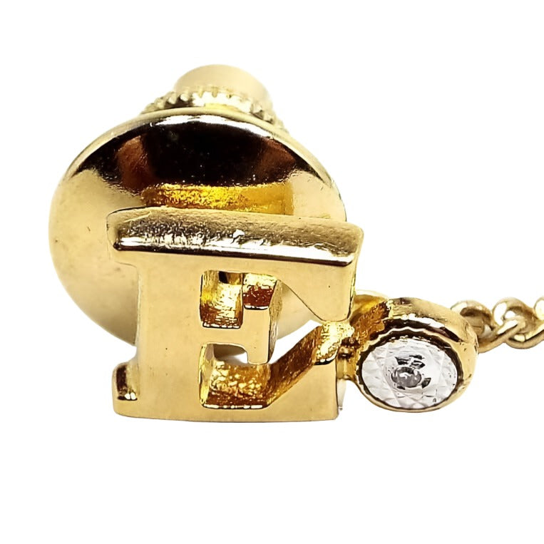 Enlarged front view of the retro vintage initial tie tack with diamond chip. The metal is gold tone plated in color. The tie tack is shaped like a block letter E. To the right of the letter is a small round area with a tiny diamond chip in it surrounded by faceted silver tone metal.