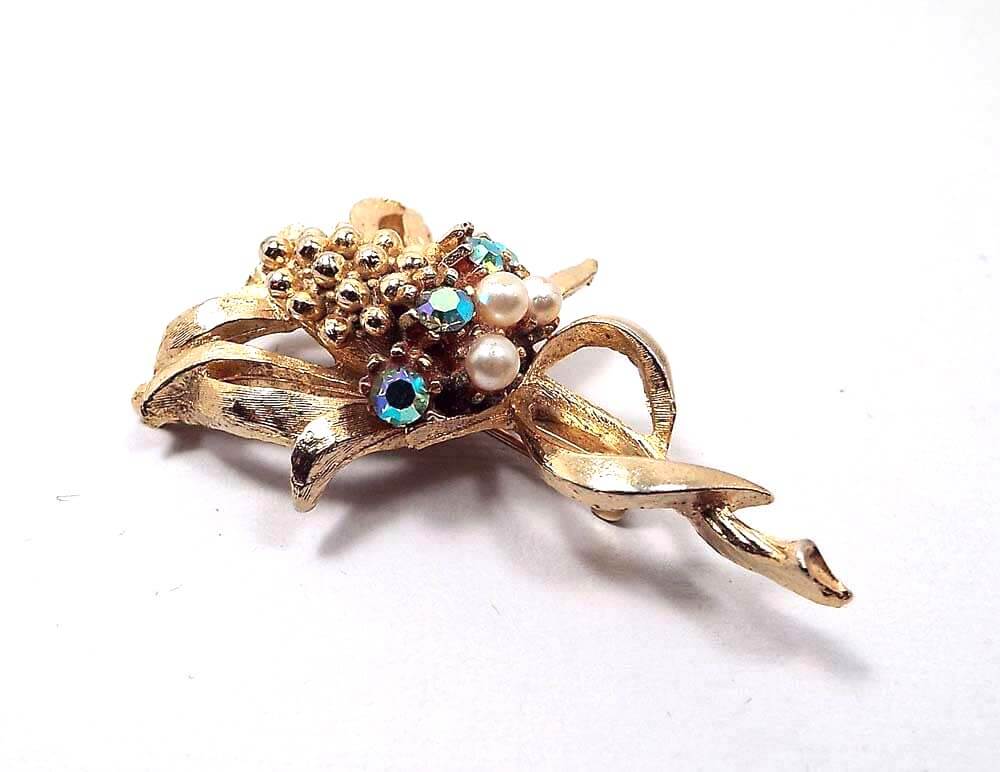 Brooch is gold in color. It looks like a corn stalk with leaves around the stalk on the bottom and curled away from the corn style piece in the middle. Under the corn are AB rhinestones that have flashes of different colors as you move, and under that is a row of imitation pearls.