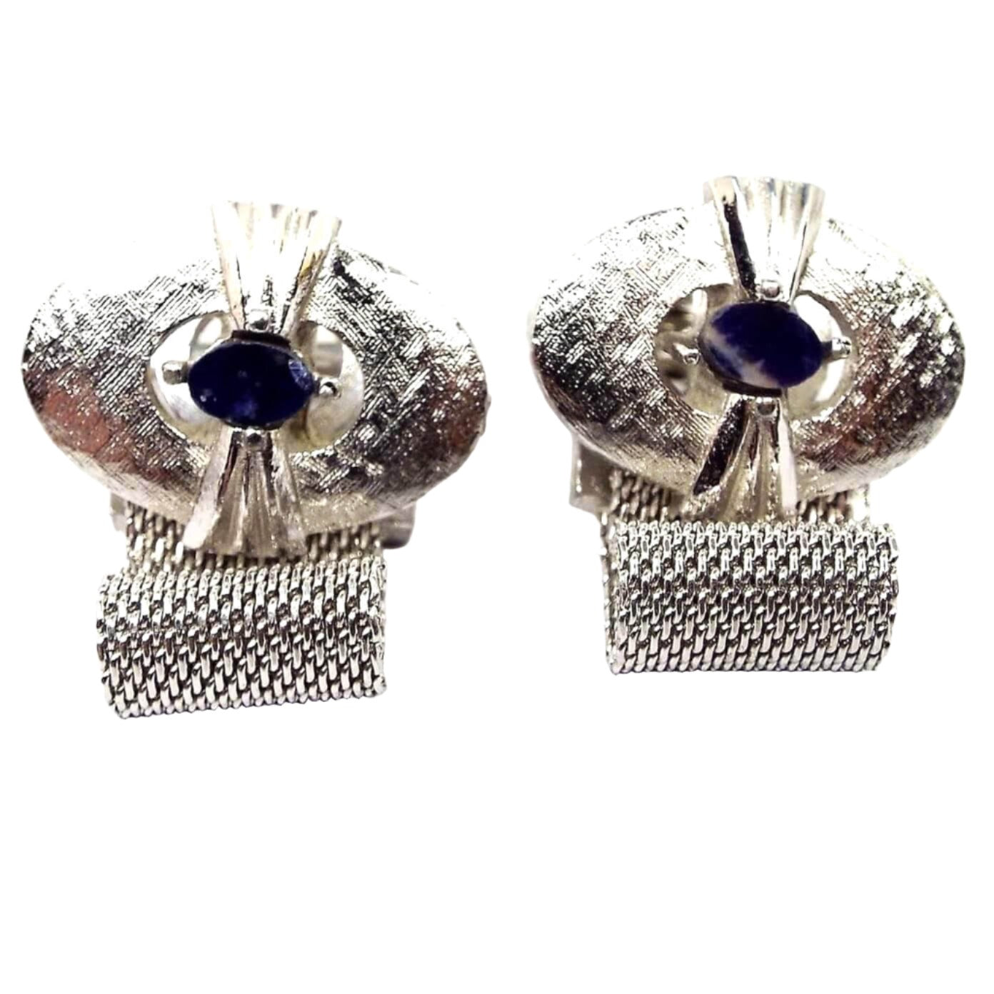 Front view of the Mid Century vintage Dante mesh cufflinks. These are larger sized. The tops have brushed matte silver tone ovals. In the middle are small oval blue sodalite gemstone cabs that have shiny silver tone metal flared out from the sides over the larger matte oval. The bottoms have mesh chain that is wide and wraps around to the back of the cufflinks. The sodalite cabs are mostly dark denim blue with a few splashes of white.
