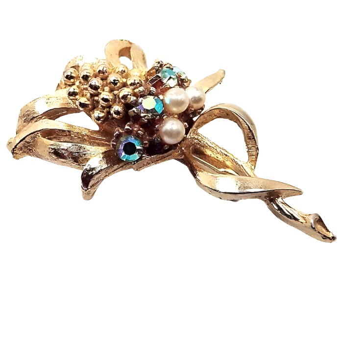 Brooch is gold in color. It looks like a corn stalk with leaves around the stalk on the bottom and curled away from the corn style piece in the middle. Under the corn are AB rhinestones that have flashes of different colors as you move, and under that is a row of imitation pearls.